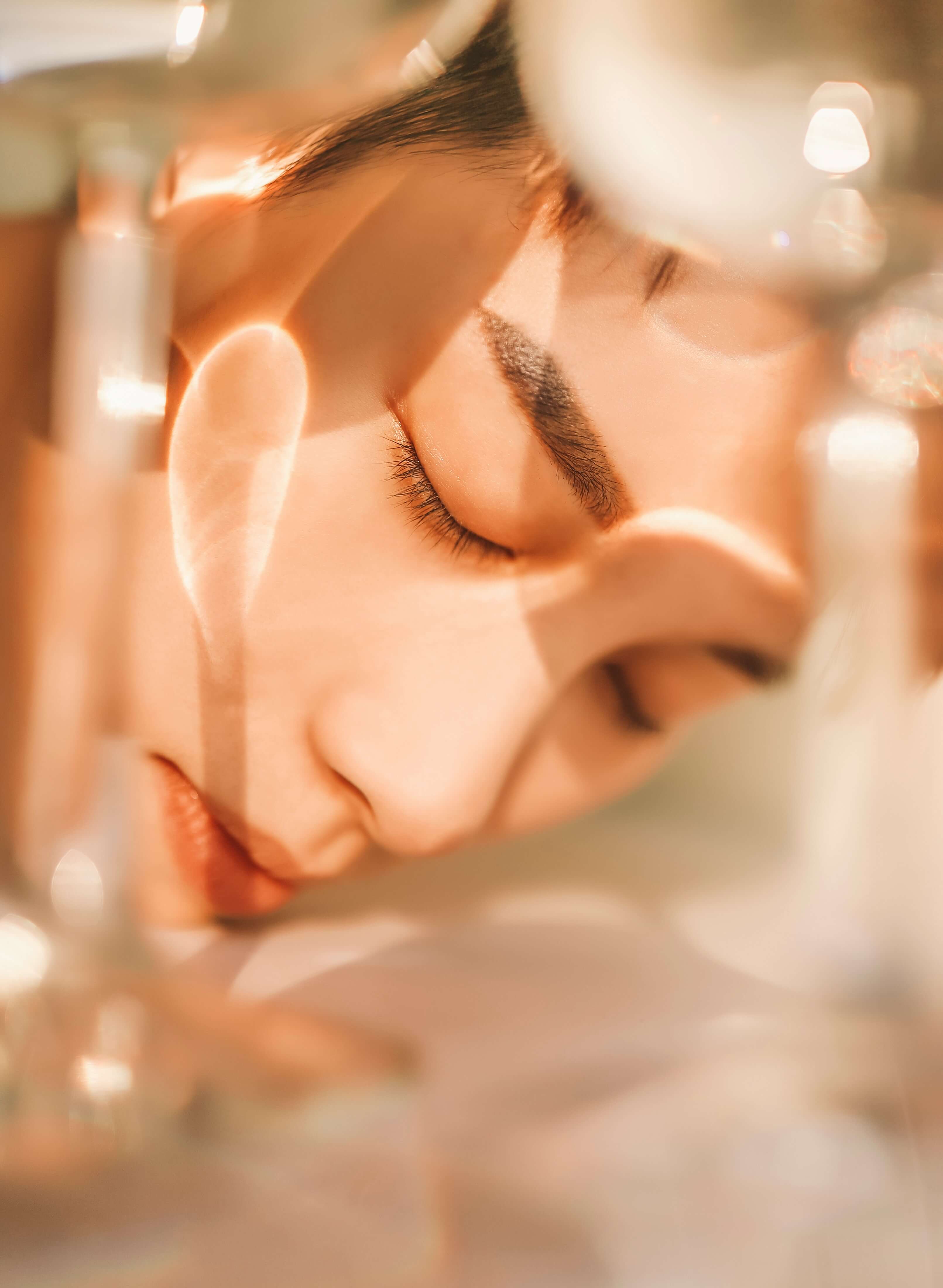 Primo piano artistico di un volto femminile immerso in riflessi luminosi e sfocature calde che richiamano il concetto di benessere, skincare e longevità cutanea.